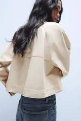 Trench corta de color beige, con cuello solapa, manga larga y cierre frontal con botones oscuros. Presenta bolsillos de vivo en el delantero y un diseño de corte recto.