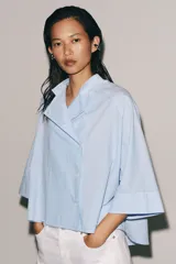 Camisa Zara Woman Collection de algodón con estampado de rayas verticales blancas y celestes. Presenta cuello perkins, mangas tres cuartos y un diseño asimétrico con cierre frontal cruzado mediante botones.