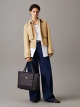 Cartera tipo shopper mediana de Calvin Klein, color negro, confeccionada en material textil con textura de lino tejido. Presenta asas fijas para el hombro, cierre de cremallera superior, bolsillo exterior frontal con cierre giratorio y herraje con monograma CK.