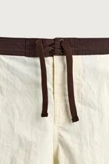 Short de baño blanco marfil con cintura marrón y cordón ajustable del mismo color. Presenta franjas laterales en tono azul marino con finos bordes rojizos y bolsillos laterales.