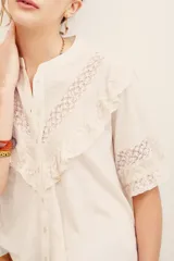 Camisa de algodón color crudo, con cuello mao, cierre frontal abotonado y mangas cortas. Presenta aplicaciones de encaje y volados en el pecho y las mangas.