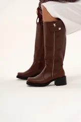 Bota de caña alta estilo biker confeccionada en cuero color chocolate. Presenta un cierre metálico lateral que recorre toda la caña, taco cuadrado y plataforma. Incluye un sistema de estribo y cadena metálica desmontable en la zona del tobillo, permitiendo diversas formas de uso.