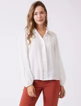 Camisa de gasa blanca con cuello solapa y cierre frontal de botones. Presenta un delicado galón de encaje festoneado a lo largo de la cartera frontal. Sus mangas largas son ligeramente abullonadas, con insertos de puntilla horizontal y puños elásticos fruncidos.