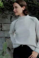 Buzo de punto color blanco con cuello alto y detalles de pespuntes decorativos en color negro en las costuras de los hombros, puños y ruedo.