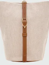 Cartera tipo bucket de tamaño pequeño, confeccionada en lona color crudo con detalles en cuero color tostado. Presenta una base redondeada, cierre de solapa con broche y correa ajustable para llevar al hombro.