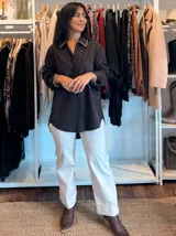 Camisa blanca de corte clásico y diseño holgado, con cuello camisero que presenta un detalle de pedrería o brillo sutil en los bordes. Posee mangas largas con puños rectos y un largo asimétrico, siendo más corta en la parte delantera y más larga en la parte trasera.