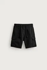 Bermuda cargo color negro de corte relaxed fit, confeccionada en tejido de algodón con elasticidad y efecto lavado. Cuenta con cintura elástica ajustable con cordón, bolsillos frontales y bolsillos laterales tipo cargo con solapa en las perneras.