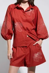 Camisa roja con bordados florales blancos, cuello camisero, cierre frontal oculto y mangas largas abullonadas.