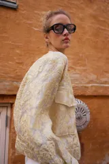 Chaqueta tipo bomber de cuello redondo y manga larga, confeccionada en tejido jacquard con diseño floral en tonos dorados y crema. Presenta cierre frontal con cremallera, bolsillos delanteros de plastrón con solapa y puños y bajo elásticos.