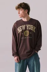 Remera de manga larga en color marrón Kahlúa, de corte holgado y cuello redondo. Presenta un estampado gráfico frontal de estilo vintage con el texto "NEW YORK", "19 75", "CHAMPIONS" y un emblema circular central.