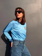 Remera de manga larga en color celeste con estampado de soles y flores de ceibo en tono azul. Presenta cuello polo y puños en tejido rib azul marino, con un parche circular bordado en el pecho que incluye el sol y la inscripción Uruguay Vdamiani.