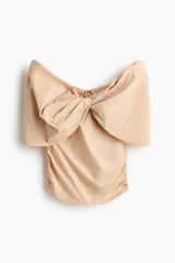 Top color beige claro, de hombros descubiertos, con amplio escote vuelto y detalle de nudo drapeado en el frente. Presenta frunces laterales y cierre posterior oculto.