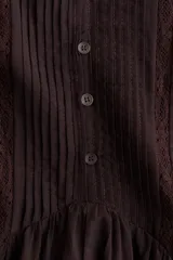 Vestido corto de color bordó, con escote en V y cuello de volados, mangas cortas abullonadas y botonadura frontal con alforzas e inserciones de encaje. Presenta costuras angulares con frunces en la cadera y falda con ribete de volado en el bajo.