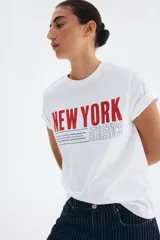 Remera blanca de manga corta y cuello redondo, con estampado frontal que dice "NEW YORK" en rojo y "ATHLETICS" en contorno negro, acompañado de un texto pequeño.