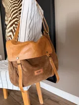 Bandolera tipo messenger bag confeccionada artesanalmente en combinación de cuero y gamuza marrón. Cuenta con solapa frontal de cuero con broches a presión, correas decorativas con hebillas metálicas y una correa larga ajustable para el hombro. Incluye parche con logo grabado en la parte frontal.