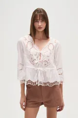 Blusa blanca de algodón con bordados tipo broderie anglaise, escote en V con lazos ajustables en el centro del busto y peplum con terminación festoneada. Mangas tres cuartos.