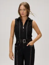 Enterito de denim negro sin mangas, con cuello camisero, cierre frontal completo y detalles de tachas metálicas a lo largo de las costuras. Incluye un cinturón ajustable con hebilla metálica y bolsillos en el frente.