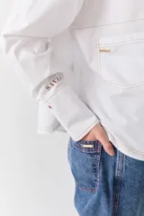 Camisa de corte recto y holgado, color blanco, con cuello mao y cierre frontal de botones. Presenta costuras a la vista en color contrastante, bolsillo lateral tipo parche y puños anchos con detalle bordado.