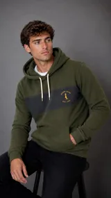 Hoodie de color verde oliva con una franja horizontal en color gris oscuro en el pecho. Presenta capucha con cordones ajustables en color crema, bolsillo tipo canguro frontal y logo bordado de la marca Duxon Jeans en color amarillo sobre la franja gris.