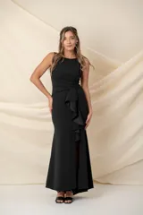 Vestido largo de fiesta color negro, confeccionado en crepé. Presenta escote redondo, corte ajustado al cuerpo, drapeado en la cintura con detalle de moño y volado vertical. Tiene tajo en la parte delantera.