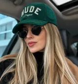 Gorra verde botella de estilo varsity, estructurada, con la inscripción frontal "NUDE Project" bordada en blanco. Presenta detalles de hojas bordadas en tono sobre tono sobre la visera.
