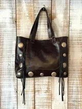 Cartera tipo tote de cuero marrón oscuro, con diseño estilo boho. Presenta apliques metálicos circulares plateados en los laterales y la base, junto con flecos de cuero negro que cuelgan de los costados. Cuenta con doble asa superior para llevar al hombro.