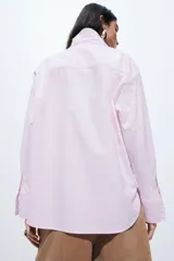 Camisa de corte holgado confeccionada en popelina de algodón, con cuello clásico, cierre frontal de botones, hombros caídos y bolsillo de parche en el pecho. Presenta mangas largas con puños abotonados, canesú en la espalda y bajo redondeado con aberturas laterales, siendo ligeramente más larga en la parte posterior.