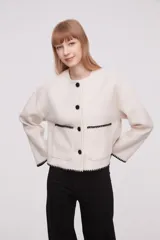 Chaqueta corta de paño color marfil, con cuello redondo, cierre frontal de botones negros y dos bolsillos frontales. Presenta detalles de costuras decorativas en contraste negro en los bordes de los bolsillos, puños y ruedo.