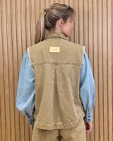 Chaleco de denim color marrón, confeccionado en 100% algodón, con bolsillos frontales amplios, cierre de botones y costuras decorativas bordadas en blanco en los bordes y los bolsillos.