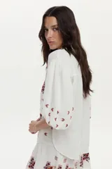 Blusa blanca de gasa de algodón con mangas 3/4 abullonadas y escote en V con botones. Cuenta con bordados en tonos marrones y naranjas en la zona frontal y las mangas.