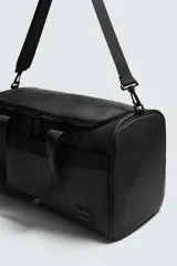 Bolso de viaje tipo duffel bag, color negro, con acabado engomado y costuras visibles. Cuenta con dos asas de mano y una correa de hombro ajustable y extraíble. Posee múltiples compartimentos con cierre de cremallera.