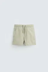Bañador de hombre color verde claro con finas rayas blancas verticales (efecto seersucker). Tiene cintura elástica ajustable con cordón verde, bolsillos laterales y un bolsillo trasero de vivo.