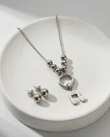 Set de joyería en acero quirúrgico compuesto por un collar con dije de nota musical y aros tipo bolita. El collar presenta una cadena con eslabones finos y una sección de esferas metálicas que conectan con un aro central del cual cuelga el dije. Los aros son de diseño minimalista en forma de esfera con cierre de presión.