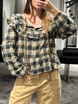 Camisa de manga larga con estampado a cuadros en tonos gris, negro y mostaza. Presenta volados en la parte superior delantera y en los hombros, con un diseño de corte holgado y lazos decorativos en el cuello.
