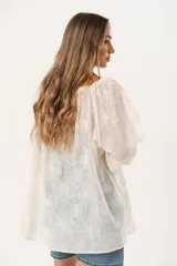 Blusa oversize de color crudo con escote bote y lazo regulable. Confeccionada en tela liviana y traslúcida con bordado floral. Presenta mangas tres cuartos con puños elásticos.