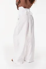 Pantalón de lino blanco de corte ancho y fluido, con cintura alta que incorpora una sobrepretina ancha desmontable con pliegues horizontales.