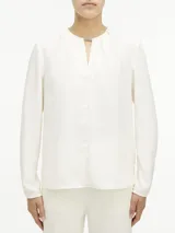 Blusa blanca de poliéster reciclado, con cuello redondo, mangas largas y abertura en el cuello con barra metálica.