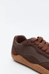 Zapatilla de estilo barefoot color marrón, confeccionada con una combinación de gamuza y materiales sintéticos. Presenta un diseño de perfil bajo con suela de goma flexible en color caramelo que incluye el logo de la marca grabado en el lateral.