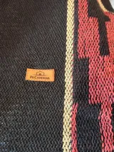 Poncho negro de hilo de algodón, con diseño geométrico tejido en tonos bordó y beige, detalles en cuerina marrón en el cuello y flecos en el ruedo.