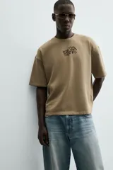 Sudadera de manga corta color beige, con cuello redondo y corte relajado. Presenta un bordado y estampado en el pecho con texto y un diseño floral.