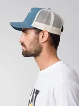 Gorro tipo trucker de color azul denim en la parte frontal y visera, con malla trasera blanca. Presenta un estampado frontal con la inscripción "Caterpillar" y la silueta de un vaquero montando un caballo. Incluye cierre trasero ajustable.