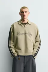 Sudadera de corte boxy fit color verde oliva, con cuello solapa y abertura frontal. Presenta mangas largas con bandas laterales negras a contraste y bordado frontal con texto en color negro.
