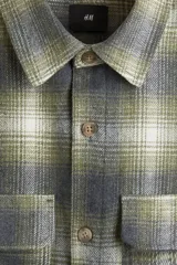 Sobrecamisa de franela suave con cuello inglés, bolsillos superiores con solapa y cierre frontal de botones. Presenta un diseño de cuadros en tonos verdes, grises y blancos, con mangas largas y bajo recto.