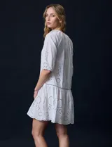 Vestido corto blanco de algodón con bordado calado tipo broderie, cuello redondo, escote en V con lazo y borlas, y mangas tres cuartos con puño.