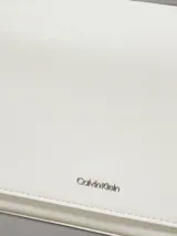 Cartera de hombro rectangular color blanco, con solapa frontal y logo metálico de Calvin Klein. Cuenta con dos correas desmontables: una fina ajustable y una ancha de tela con inscripción.