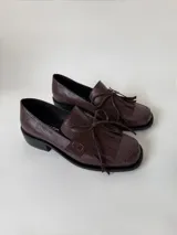 Mocasín de cuero marrón con textura granulada, diseño de horma cuadrada, detalle de flecos y lazo decorativo en el empeine, montado sobre una suela y taco bajo de color negro.