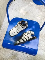 Sandalias planas de cuero color blanco hueso, con múltiples tiras horizontales y hebillas metálicas plateadas. Incluyen pulsera superior desmontable con hebillas laterales para regular el ajuste.