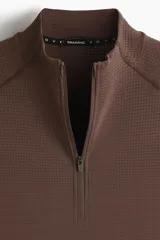 Remera deportiva marrón con cuello alto, cierre frontal parcial y mangas largas raglán, confeccionada en tejido técnico con paneles texturizados y corte ajustado.