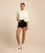 Short de sarga color blanco, de cintura alta, con bolsillos delanteros y traseros, trabillas para cinturón y bajo plegado. Cuenta con cierre frontal de botón y cremallera.