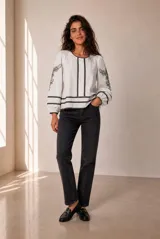 Blusa blanca de manga larga con detalles de bordados florales en tono grisáceo y bandas decorativas en el frente y mangas. Cuenta con cuello redondo y puños elásticos.